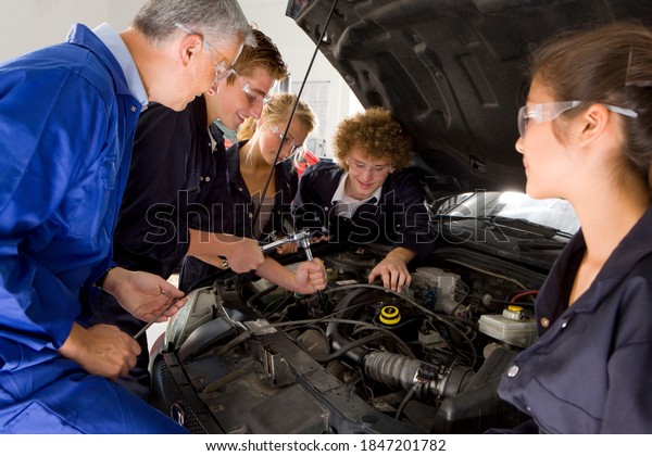 Young students leaning\
under the hood of a car in order to repair it with their trainer\
standing beside as he carefully observes them working in the\
automotive training\
school
