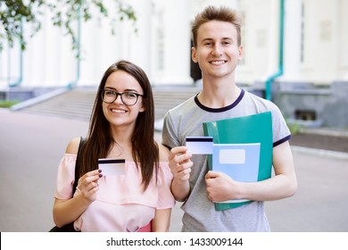 Young Students Near University With Books, Exercise Books Holding Credit Cards And Smiling To Camera. Modern College Learners Using Cards For Online Banking, Buying Things Online, Receiving Stipend