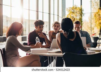 Young Students Sitting Around A Table In Library With Books And Laptop. University Students Doing Group Study In Library.