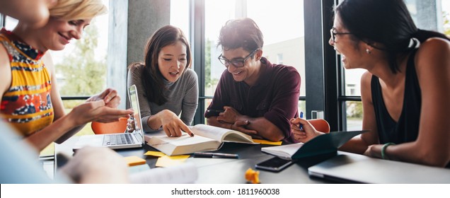 Young Students Sitting At A Table In A Library And Reading Books For Their Class Assignment. University Students Doing Group Study In Library.