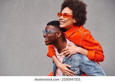 Young and stylish african american woman in sunglasses piggybacking on best friend in denim jacket and having fun near building on urban street, trendy friends in urban settings - Powered by Shutterstock