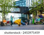 Young Stylish B-boy Breakdancing On City Street Among Buildings In Urban Area. Fashionable Group Of Friends Chilling On Background, Supporting Breakdance Performer Practising Choreography.