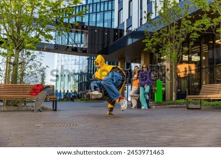 Young Stylish B-boy Breakdancing On City Street Among Modern Buildings In Urban Area. Fashionable Group Of Friends Relaxing On Background, Supporting Breakdance Performer Practising Choreography.