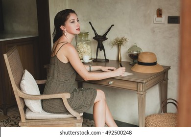 Young Stylish Beautiful Woman Sitting At Table In Resort Hotel Room, Writing A Letter, Thinking, Sophisticated, Smiling, Happy, Bohemian Outfit, Holding Pen, Straw Hat, Vintage Style