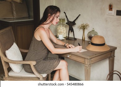 Young Stylish Beautiful Woman Sitting At Table In Resort Hotel Room, Writing A Letter, Thinking, Sophisticated, Smiling, Happy, Bohemian Outfit, Holding Pen, Straw Hat, Vintage Style