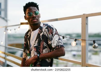 Young Stylish Black Man In Casualwear And Sunglasses Holding Bottle Of Beer While Standing Against Urban Environment In Rooftop Cafe