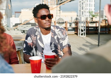 Young Stylish Black Man In Sunglasses And Casualwear Having Drink In Outdoor Cafe While Sitting By Table Among His Friends