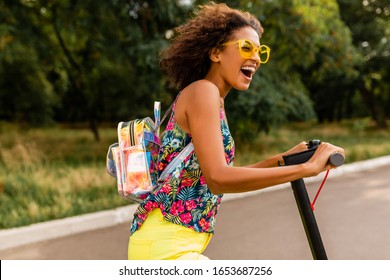 young stylish black woman having fun in park riding on electric kick scooter in summer fashion style, colorful hipster outfit, wearing backpack and yellow sunglasses - Powered by Shutterstock