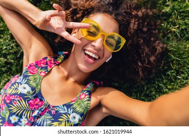young stylish black woman listening to music on wireless earphones having fun in park, summer fashion style, colorful hipster outfit, lying on grass, african hairstyle, view from above