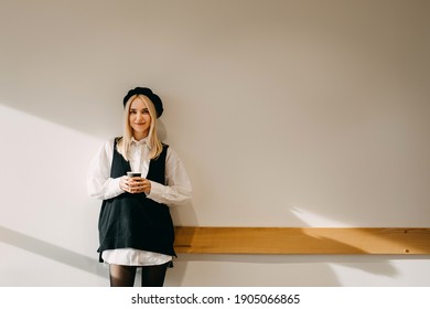 Young stylish blonde woman standing against a white wall, holding a cup of coffee, smiling.