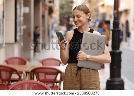 Young stylish business woman freelance is holding laptop and using smartphone in european city street. Concept of remote work or leisure time with gadgets outdoors. Idea of modern communication.