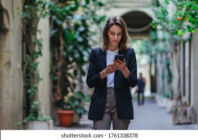 Young Stylish Business Woman Writes Message On Smartphone Walking On Old City Street, Hipster Tourist Traveling In European Town And Planning Trip Using Mobile Phone 