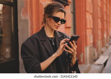 Young Stylish Business Woman Writes Message On Smartphone Walking On Old City Street, Hipster Tourist Traveling In European Town And Planning Trip Using Mobile Phone.