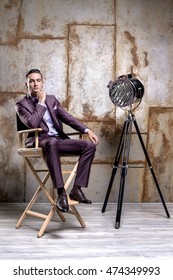 Young Stylish Businessman In Fashionable Suit Posing In The Photo Studio . Luxury Portrait Of A Man Sitting In The Director's Chair Next To The Model Is A Retro Movie Spotlight