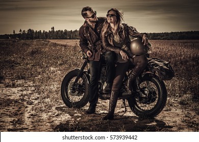 Young, stylish cafe racer couple on vintage custom motorcycles in field.