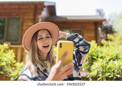 Young Stylish Caucasian Woman 30-35 Years Old Wearing Casual Clothing Shirt And Cowboy Hat. Influencer Using Social Media Via Mobile Phone. Person Enjoys Life At Morning During Summer Adventure In