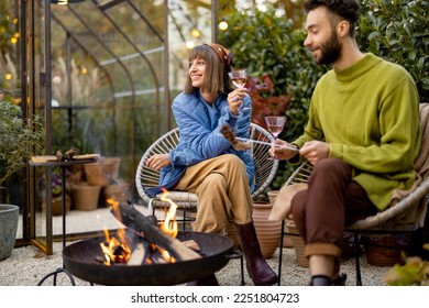 Young stylish couple grilling food and warming up while sitting together by the fire, spending autumn evening time at cozy atmosphere in garden - Powered by Shutterstock