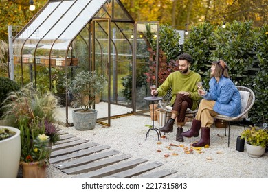 Young Stylish Couple Hang Out Together, Sitting With Wine By The Fire, Spending Evening Time At Cozy Atmosphere In Beautiful Backyard With Greenhouse On Background