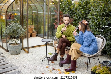 Young Stylish Couple Hang Out Together, Sitting With Wine By The Fire, Spending Evening Time At Cozy Atmosphere In Beautiful Backyard With Greenhouse On Background