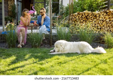 Young Stylish Couple Have A Drink While Sitting Together With Dog On A Lawn At Their Beautiful Backyard Of Country House. Young Man And Woman Spend Summer Time