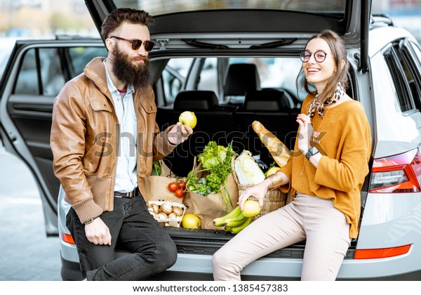 Young stylish couple having fun while sitting on\
the car trunk full of fresh and healthy food on the supermarket\
parking outdoors