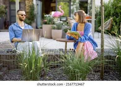 Young Stylish Couple Work Or Spend Leisure Time With Laptop Computer And Digital Tablet On Porch Of Their Country House. Concept Of Remote Work At Cozy Home Atmosphere