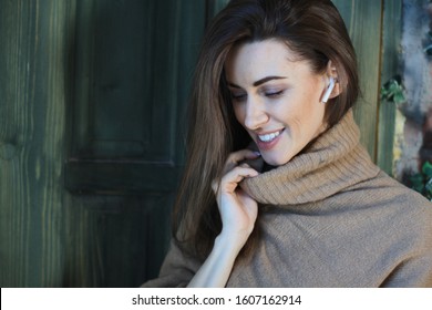 Young Stylish Fashion Straight Brown Hair Smile Laughing Face Woman In Winter Oversize Sweater Talks By Phone With Wireless Earphones With Nature Wooden House Door In Cozy Holiday House Interior