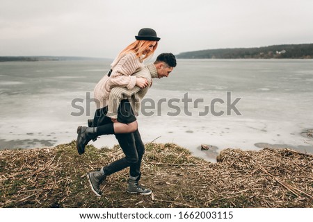 Young stylish loving couple on the shore of a frozen lake. The girl sits on her back with a boyfriend, happiness and fun. Multicultural couple, asian and european with red hair and black hat.