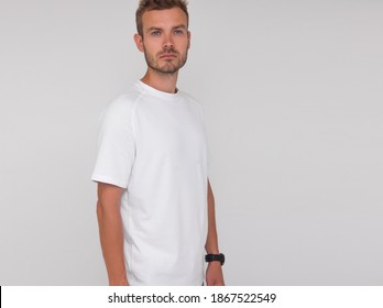 Young Stylish Male Doctor In White T-shirt Is Standing In Profile And Smiling On The White Wall Background. Medical Concept. Free Space On Right Side, Mockup	
