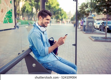 Young stylish man with headphones sitting on bus stop and typing sms