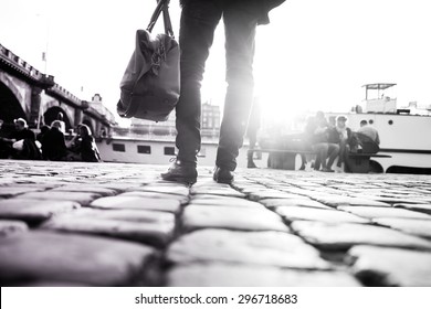 Young Stylish Man With His Bag Is Standing In Front Of River And Looking In The City. Detail Of His Shoes And His Bag. Moment Of The Arriving The City. Black And White Photography.