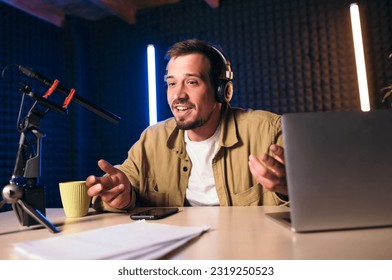 Young stylish man in mustard shirt with headphones gesturing at microphone and sharing story with audience while sitting at desk in studio with neon lighting and recording podcast