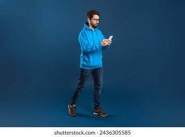 Young Stylish Man Walking With Smartphone In Hands Over Blue Studio Background, Handsome Millennial Guy Messaging With Friends On Mobile Phone Or Shopping Online, Full Length With Copy Space - Powered by Shutterstock