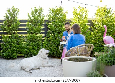 Young Stylish Man And Woman Sit Relaxed With A Dog And Have A Dinner At Beautiful Garden Of Country House. Young Family Spending Summer Time At Backyard