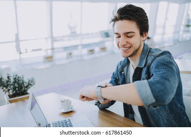 Young Stylish Man Working On A Macbook Or Laptop In Cafe While Having Coffee And Checking His Nice Watch. Happy Guy In Jeans Jacket Pointing At Clock Face