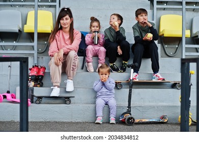 Young Stylish Mother With Four Kids Sitting On The Sports Podium At The Stadium, Eat Apple And Drink Water. Family Spend Free Time Outdoors With Scooters And Skates.