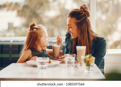 young and stylish mother with long hair and a green dress sitting with her little cute daughter in the summer cafe and she feeds her daughter with a dessert - Powered by Shutterstock