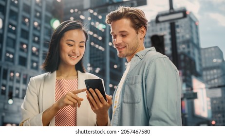 Young Stylish Multiethnic Couple is Casually Walking on a Street in a Big City. Attractive Japanese Female Showing Smartphone to Handsome Caucasian Male. Diverse Friends Enjoying Travelling Together.