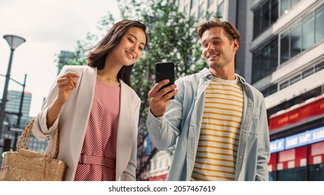 Young Stylish Multiethnic Couple is Casually Walking on a Street in a Big City. Handsome Caucasian Male Showing Smartphone to Beautiful Japanese Female. Diverse Friends Enjoying Travelling Together. - Powered by Shutterstock