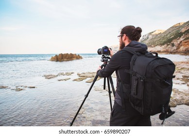 Young stylish photographer with beard and backpack making photos of sea and rocks with the camera on a tripod. Mediterranean sea - Powered by Shutterstock