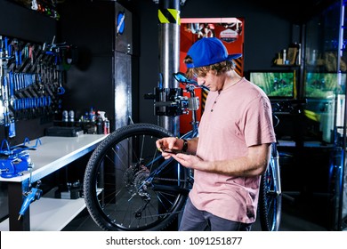 A young, stylish redheaded male small business owner selling and repairing a bike is wearing a blue cap and a pink jersey is using a mobile phone while standing in a bike shop. - Powered by Shutterstock