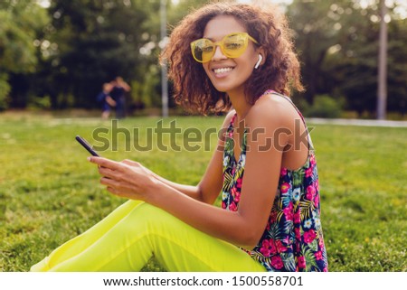 young stylish smiling black woman using smartphone listening to music on wireless earphones having fun in park, summer fashion colorful style, sitting on grass, yellow sunglasses, african hairstyle
