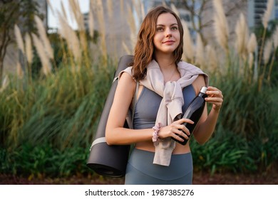 Junge stylische Sportlerin, die im Sommer draußen steht und eine Flasche Wasser und Yogamat hält.