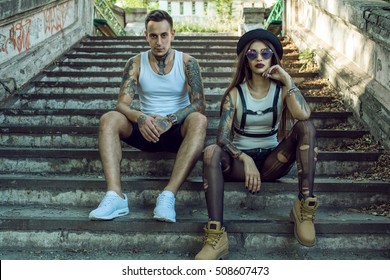 Young stylish tattooed couple in black shorts sitting on the ruined stairs and looking straight. Girl wearing hat, torn tights and mirrored sunglasses. Grunge style concept. Outdoor shot