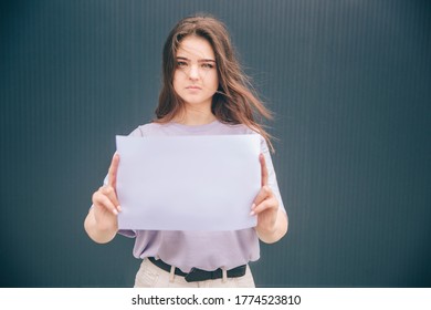 Young Stylish Trendy Woman Isolated Over Grey Blue Background. Upset Unhappy Girl Holding Blank Page Or Piece Of Paper. Wind Blowing Her Hair.