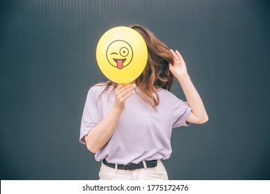 Young stylish trendy woman isolated over grey blue background. Girl hiding her face behind yellow ballon with funny emoji. Covering herself from camera.
