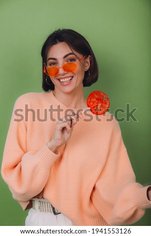 Young stylish woman in casual peach sweater and orange glasses isolated on green olive background with orange lollipop positive smile copy space