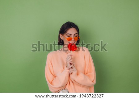 Young stylish woman in casual peach sweater and orange glasses isolated on green olive background with orange lollipop positive smile copy space