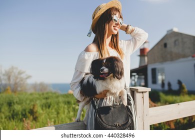 young stylish woman in countryside, holding a dog, happy positive mood, summer, straw hat, bohemian style outfit, sunglasses, smiling, happy, sunny