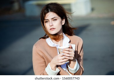 Young stylish woman drinking tea in a city street. Hipster european girl with latte paper glass  in street. Gorgeous young woman with cup of coffee in city street. Coffee break. Coffee to go. 